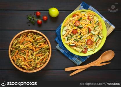 Overhead shot of raw fusilli pasta in wooden bowl and a plate of vegetarian pasta salad made of tricolor fusilli, sweet corn, cucumber and cherry tomato, photographed on dark with natural light