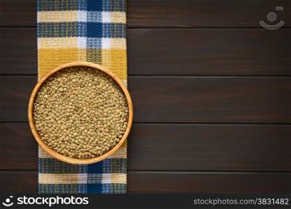 Overhead shot of raw lentils (lat. Lens culinaris) in wooden bowl, photographed on kitchen towel on dark wood with natural light