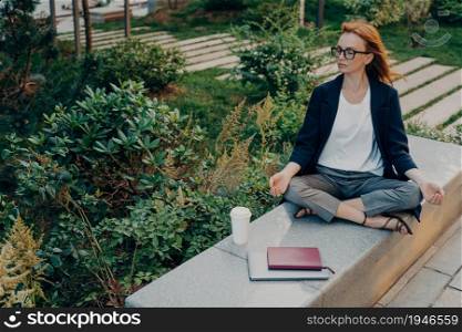 Overhead shot of relaxed ginger woman meditates in park has peaceful meditation wears glasses formal outfit drinks takeaway coffee surrounded by notepad tablet enjoys nature. Healthy lifestyle. Relaxed ginger woman meditates in park has peaceful meditation enjoys nature