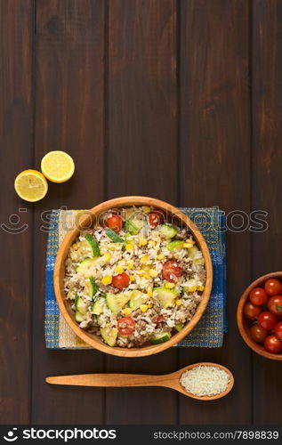 Overhead shot of rice dish with mincemeat and vegetables (sweet corn, cherry tomato, zucchini, onion) in wooden bowl with ingredients on the side, photographed on dish towel on dark wood with natural light