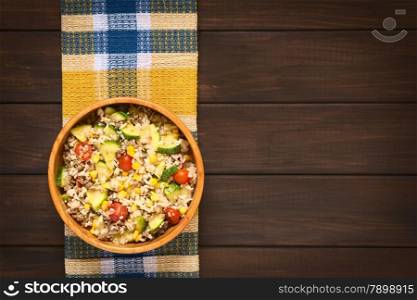 Overhead shot of rice dish with mincemeat and vegetables (sweet corn, cherry tomato, zucchini, onion) in wooden bowl, photographed on dish towel on dark wood with natural light