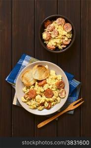 Overhead shot of scrambled eggs made with chorizo slices and onion on plate with toasted baguette slices, wooden fork on the side, photographed on dark wood with natural light