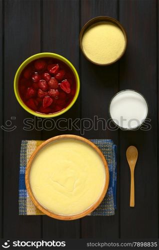 Overhead shot of semolina pudding served in wooden bowl, with a spoon, glass of milk, bowl of strawberry compote, and a bowl of uncooked semolina, photographed on dark wood with natural light