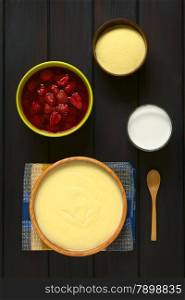 Overhead shot of semolina pudding served in wooden bowl, with a spoon, glass of milk, bowl of strawberry compote, and a bowl of uncooked semolina, photographed on dark wood with natural light
