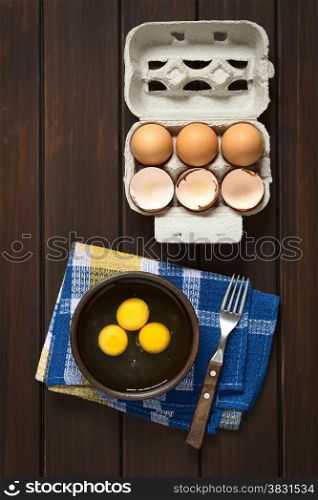 Overhead shot of three raw eggs in rustic bowl with fork on the side and egg box with eggs and eggshells above, photographed on dark wood with natural light