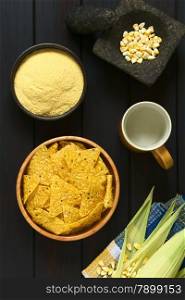 Overhead shot of tortilla chips in wooden bowl surrounded by its ingredients water, cornmeal and corn, photographed on dark wood with natural light