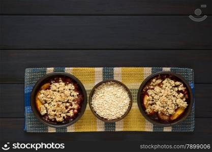 Overhead shot of two rustic bowls filled with baked plum and nectarine crumble or crisp and a bowl of rolled oats, photographed on cloth on dark wood with natural light