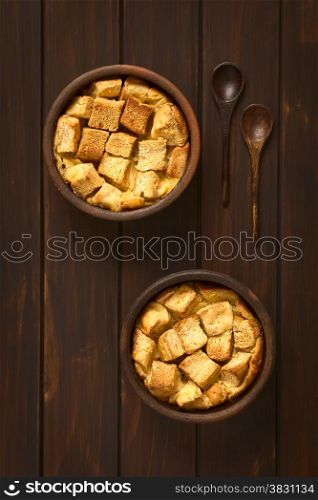 Overhead shot of two rustic bowls of bread pudding made of diced stale bread, milk, egg, cinnamon, sugar and butter, photographed on dark wood with natural light