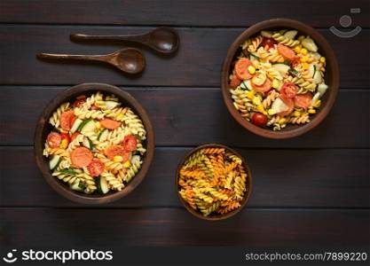 Overhead shot of two rustic bowls of pasta salad made of tricolor fusilli, sweet corn, cucumber, cherry tomato and sausage, photographed on dark wood with natural light
