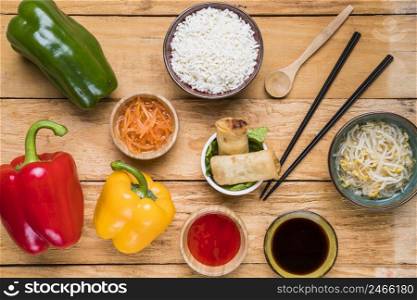 overhead view bell peppers carrot rice spring rolls sprout beans sauces with chopsticks ladle wooden desk