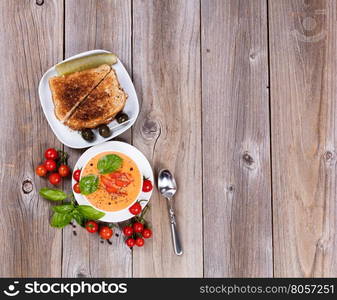 Overhead view of a bowl of fresh creamy tomato soup, tuna sandwich, with cherry tomatoes and basil leaves on rustic wooden boards.