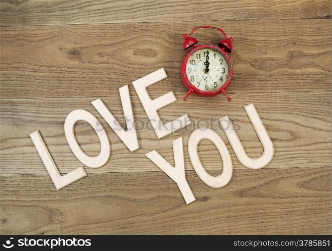 Overhead view of an old table top alarm clock and large wooden letters spelling out Love You on rustic wood