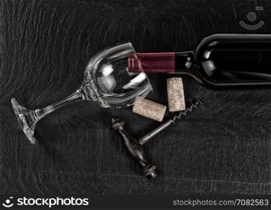 Overhead view of antique wine corkscrew, red wine bottle, glass and used corks on black slate
