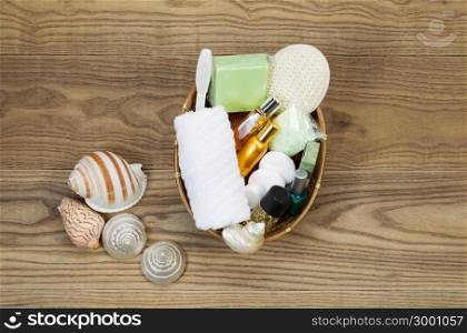 Overhead view of bath and shower accessories placed in basket on rustic wooden boards. Items include hand towel, scrub brush, lotion, soap, soaking salts, perfume, and sea shells.