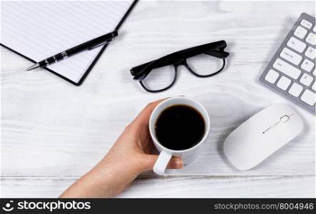 Overhead view of female hand holding cup of black coffee on white desktop with computer keyboard, mouse, reading glasses, notepad and pen.