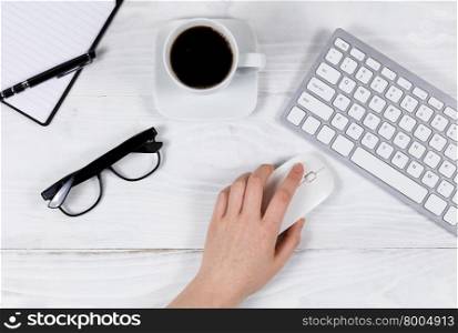 Overhead view of female hand using mouse on white desktop with computer keyboard, black coffee, reading glasses, notepad and pen.