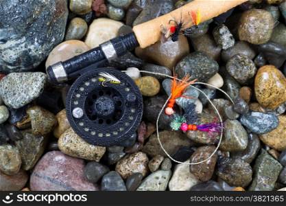 Overhead view of fishing fly reel and assorted flies on wet river bed stones