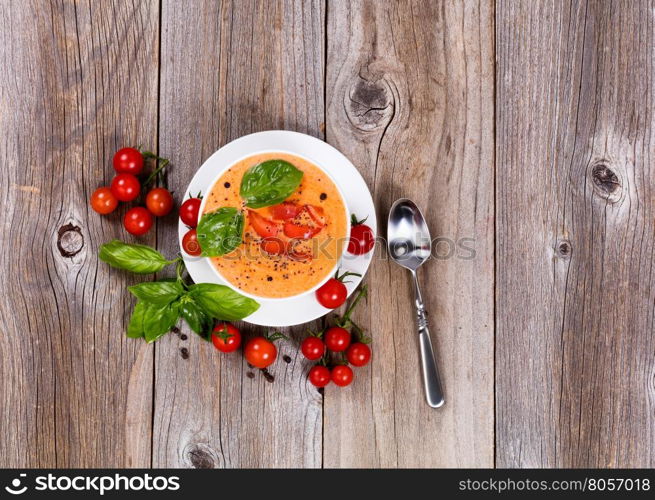 Overhead view of fresh creamy tomato soup, in white bowl, with cherry tomatoes and basil on rustic wooden boards.