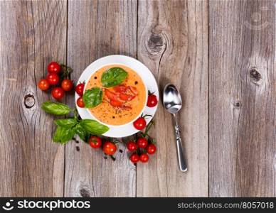 Overhead view of fresh creamy tomato soup, in white bowl, with cherry tomatoes and basil on rustic wooden boards.