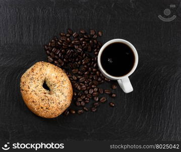 Overhead view of fresh dark coffee, large seeded bagel and roasted beans on natural slate stone.
