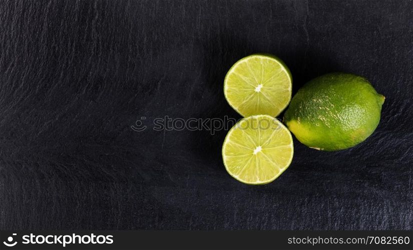 Overhead view of fresh limes, whole and sliced, on black slate with available copy space on left side