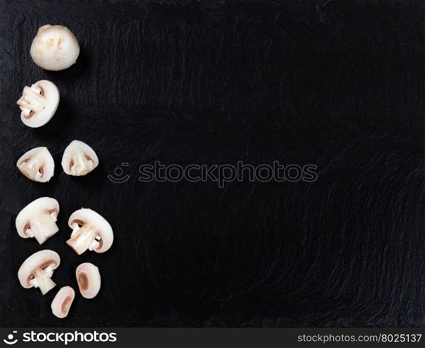 Overhead view of fresh whole and sliced mushrooms, left side of frame, on black slate.