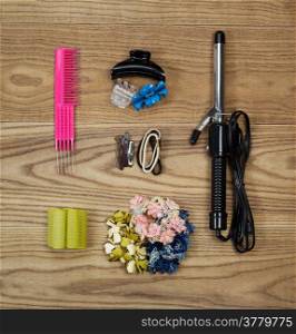 Overhead view of hair accessories placed on faded wood. Items including, curly iron, clips, rollers, elastics, hair pins, and comb.