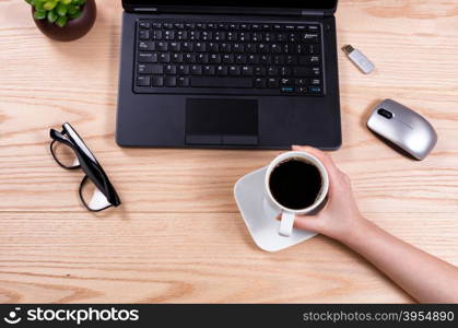 Overhead view of hand holding coffee cup with laptop, baby plant, mouse, thumb drive, and reading glasses on desktop.