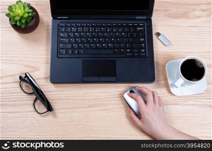 Overhead view of hand holding computer wireless mouse with laptop, baby plant, coffee, thumb drive, and reading glasses on desktop.
