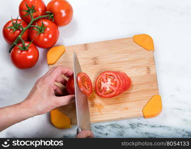 Overhead view of hand slicing a fresh garden tomato with large kitchen knife and whole tomatoes on natural bamboo cutting board.