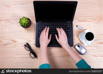 Overhead view of hands typing on laptop keyboard with wireless mouse, baby plant, coffee, thumb drive, and reading glasses on desktop.