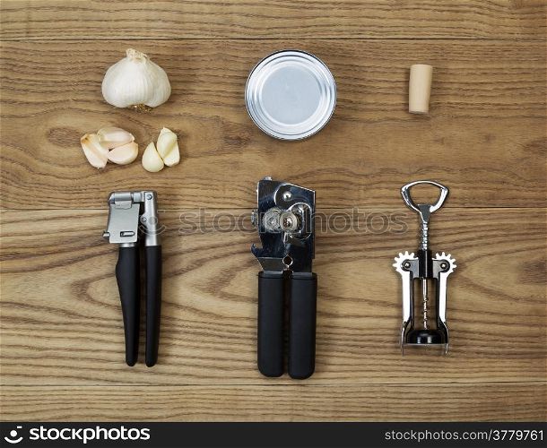Overhead view of kitchen utilities placed on rustic wood. Items including can opener, metal can, cork screw, cork, bottle opener, garlic press, and raw garlic.
