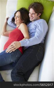 Overhead View Of Man And Pregnant Wife Watching TV On Sofa