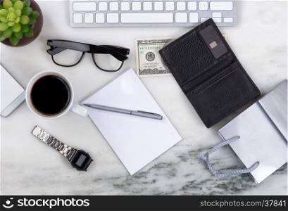 Overhead view of office desktop with computer keyboard, cell phone, shopping bag, wallet, money, pen, paper, reading glasses, coffee and plant on marble surface. Shopping on line concept.