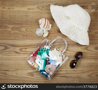 Overhead view of outdoor kit placed on rustic wooden boards. Items include clear plastic bag, comb, sun screen, hair clips, sea shells, sun glasses and white hat.