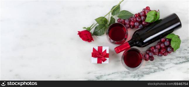 Overhead view of Red wine bottle with grapes, drinking glasses, corkscrew, gift box and rose on natural marble stone