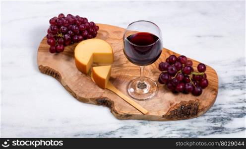 Overhead view of red wine, cheese, and grapes on wooden server with marble counter underneath. Selective focus on top of wine glass.