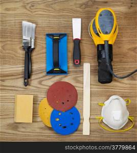Overhead view of sanding and painting equipment positioned on rustic wooden boards. Items include electric sander, mask, stir stick, sand paper, scrapper, sanding block, and brushes.