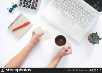 Overhead view of woman hand holding bread and cup of coffee on work space with laptop