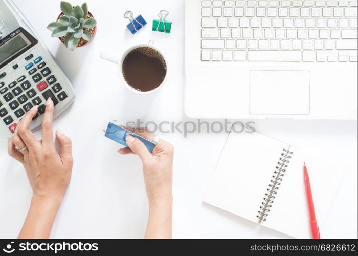 Overhead view of woman hand using calculator and holding credit card with laptop and stationery on white table