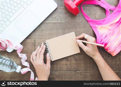 Overhead view of woman hand writing on notebook with laptop and fitness items on wooden table