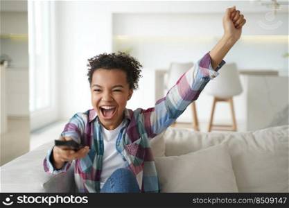 Overjoyed african american teen girl celebrating goal, sitting on couch in living room at home. Excited happy young woman watching tv football play, rooting for favorite sport team, shouting.. Overjoyed african american teen girl celebrates goal, watching television, sitting on sofa at home