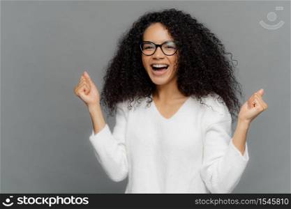 Overjoyed African American woman clenches fists with triumph, laughs with happiness, rejoices success, wears spectacles, white sweater, cheers for favourite team, isolated on grey background