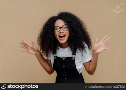 Overjoyed African American woman laughs out, raises palms, being in high spirit, wears transparent glasses, casual t shirt and overalls, poses against brown background, feels happiness and pleasure
