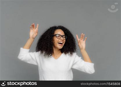 Overjoyed Afro American woman raises hands, laughs happily, enjoys pleasant music, wears optical glasses and white casual jumper, models over grey background, copy space above for your text.