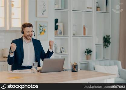 Overjoyed business professional enjoying success, raising clenched fists and screaming from happiness while using laptop and headset, sitting at his workplace at home or office. Excited successful businessman enjoying triumph with clenched fists