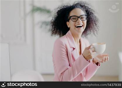 Overjoyed curly haired woman laughs happily while drinks hot coffee or latte, wears transparent spectacles, has fun during break in office, shows white teeth, being professional entrepreneur