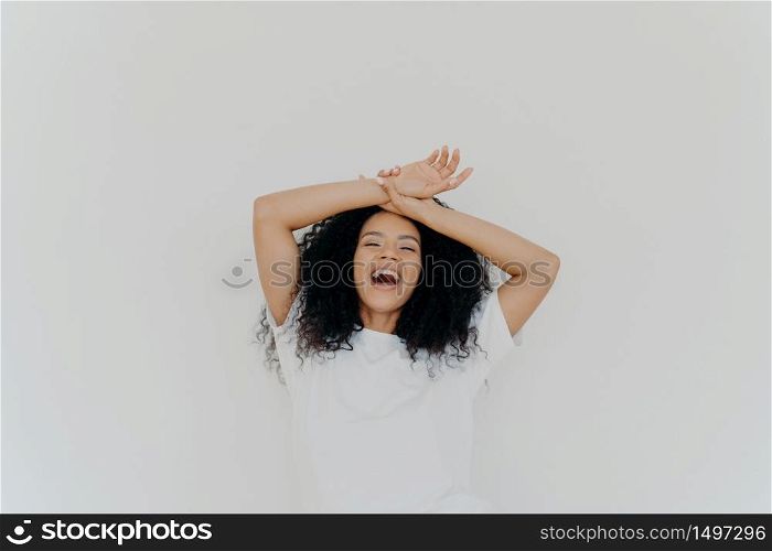 Overjoyed curly haired young lady keeps both hands on forehead, opens mouth and laughs happily, feels energetic, dressed in casual wear, poses against white studio background. What funny joke!