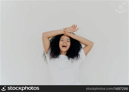 Overjoyed curly haired young lady keeps both hands on forehead, opens mouth and laughs happily, feels energetic, dressed in casual wear, poses against white studio background. What funny joke!