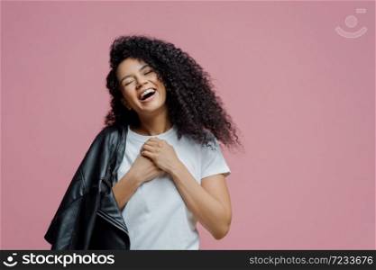 Overjoyed dark skinned woman keeps hands together and laughs sincerely, closes eyes and feels very happy, shows white perfect teeth, wears t shirt and leather jacket on shoulder, isolated on pink wall